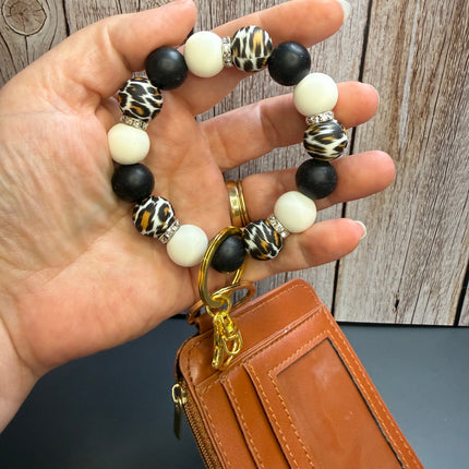 A hand holds a beaded bracelet keychain with black, white, and leopard print beads attached to a gold ring and brown leather card holder wallet, set against a rustic wood panel background.