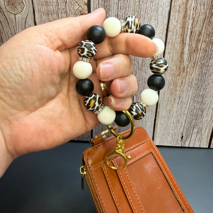A hand holds a beaded keychain bracelet with black, cream, and leopard-print beads attached to a brown leather card holder wallet with a gold key ring, set against a rustic wood panel background.