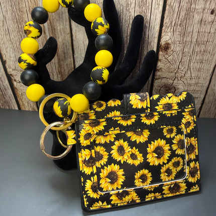 A black card holder wallet with a yellow and brown sunflower pattern, shown with a matching beaded wrist strap and gold-tone key rings on a black display mannequin hand against a rustic wood background.