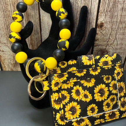 A black card holder wallet with a yellow and black sunflower pattern, displayed next to a matching beaded bracelet on a black hand-shaped stand against a rustic wood background.