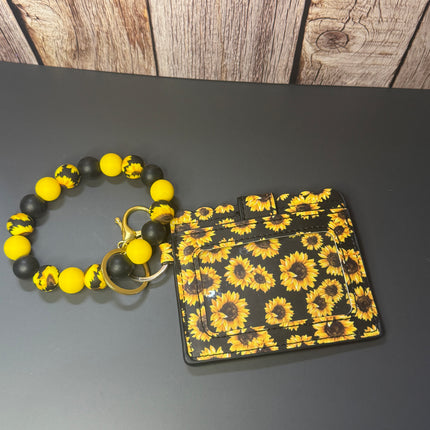 A black card holder wallet with a yellow and black sunflower pattern, attached to a matching beaded bracelet with yellow and black beads, displayed on a dark gray surface with a wood panel background.