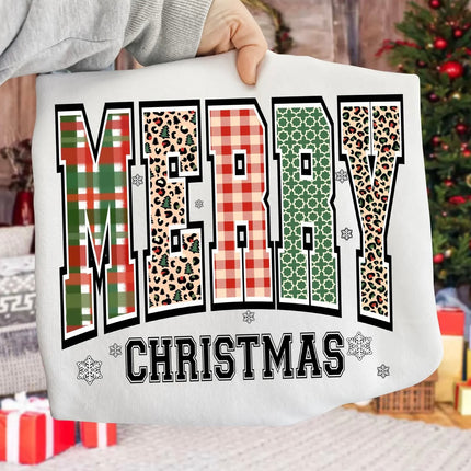 A person holds up a white sweatshirt featuring a festive graphic that reads 'MERRY CHRISTMAS' in large patterned letters. The background shows a cozy holiday setting with a Christmas tree and wrapped gifts.