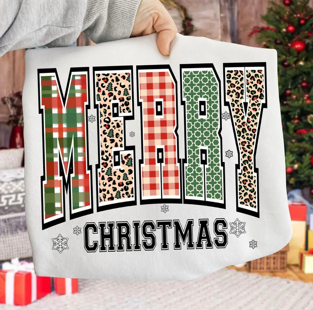 A person holds up a white sweatshirt featuring a festive graphic that reads 'MERRY CHRISTMAS' in large patterned letters. The background shows a cozy holiday setting with a Christmas tree and wrapped gifts.