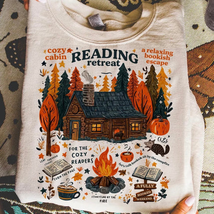 A person holds up a cream-colored crewneck sweatshirt featuring a detailed autumn-themed graphic of a cozy cabin, campfire, pumpkins, books, and the text 'READING retreat'.