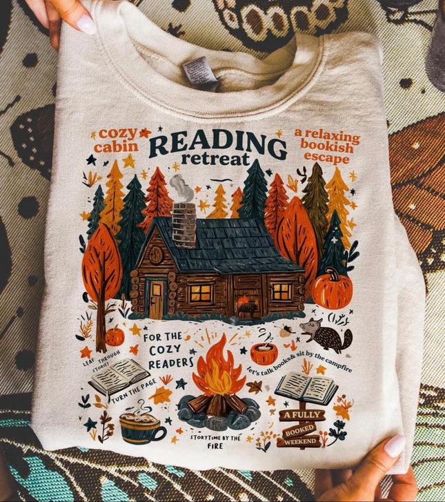 A person holds up a cream-colored crewneck sweatshirt featuring a detailed autumn-themed graphic of a cozy cabin, campfire, pumpkins, books, and the text 'READING retreat'.