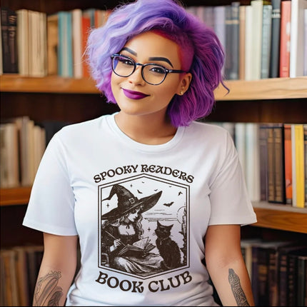 A person with purple hair and glasses wearing a white graphic T-shirt that reads 'SPOOKY READERS BOOK CLUB' with a witch and cat illustration, standing in front of bookshelves.