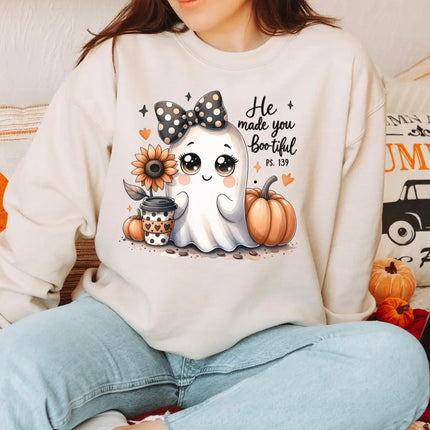 A woman wearing a cream-colored sweatshirt with a cute cartoon ghost graphic and the text 'He made you Boo hool Ps. 139' alongside pumpkins, a sunflower, and a coffee cup, paired with light blue jeans.