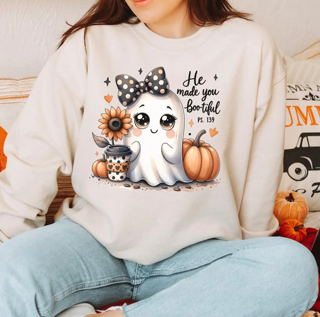 A woman wearing a cream-colored sweatshirt with a cute cartoon ghost graphic and the text 'He made you Boo hool Ps. 139' alongside pumpkins, a sunflower, and a coffee cup, paired with light blue jeans.