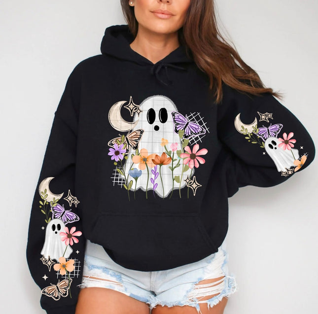 A woman wearing a black hoodie with a Halloween-themed graphic featuring a white ghost, colorful flowers, butterflies, and moons on the front and sleeves, paired with light-wash distressed denim shorts.