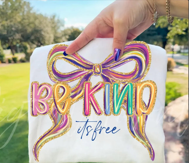 Hand holding a white shirt with a colorful bow and 'Be Kind it's free' text against a blurred outdoor background.