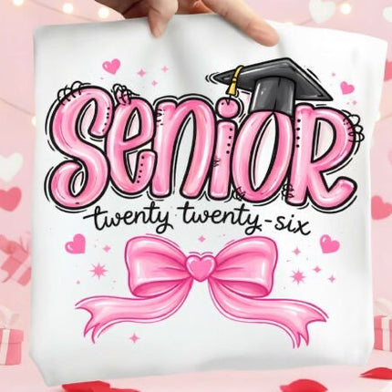 Graduation-themed pillow with 'Senior twenty twenty-six' text and pink bow on a pink heart patterned background.
